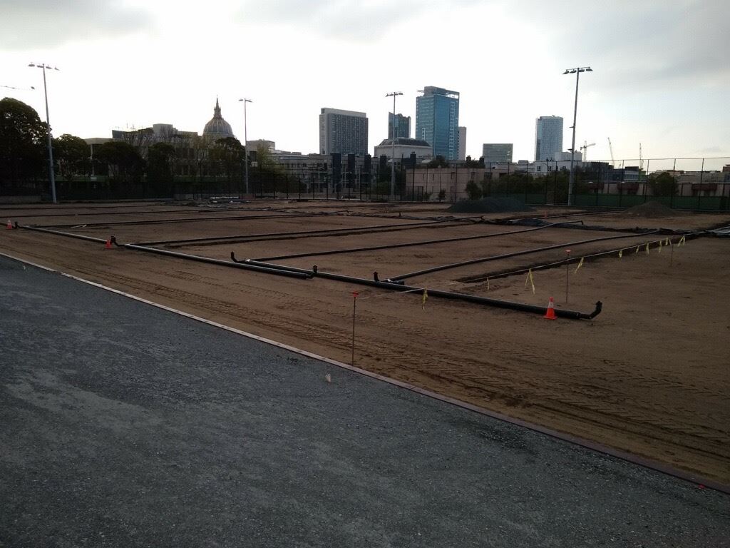 The image shows black irrigation pipes being installed in the foreground and San Francisco City Hall