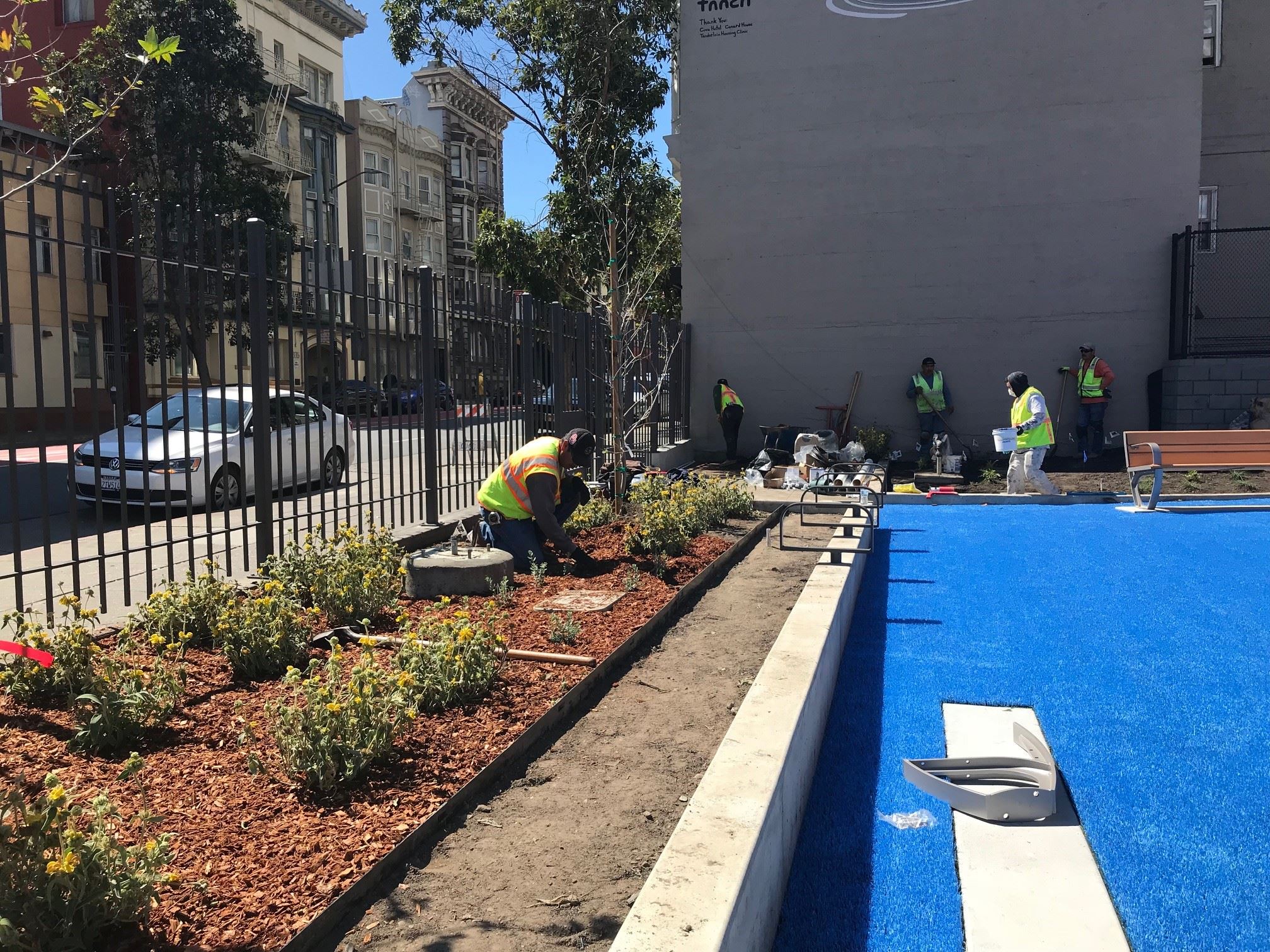 Image shows a worker wearing a yellow safety vest planting plants in a flower bed on the left side o