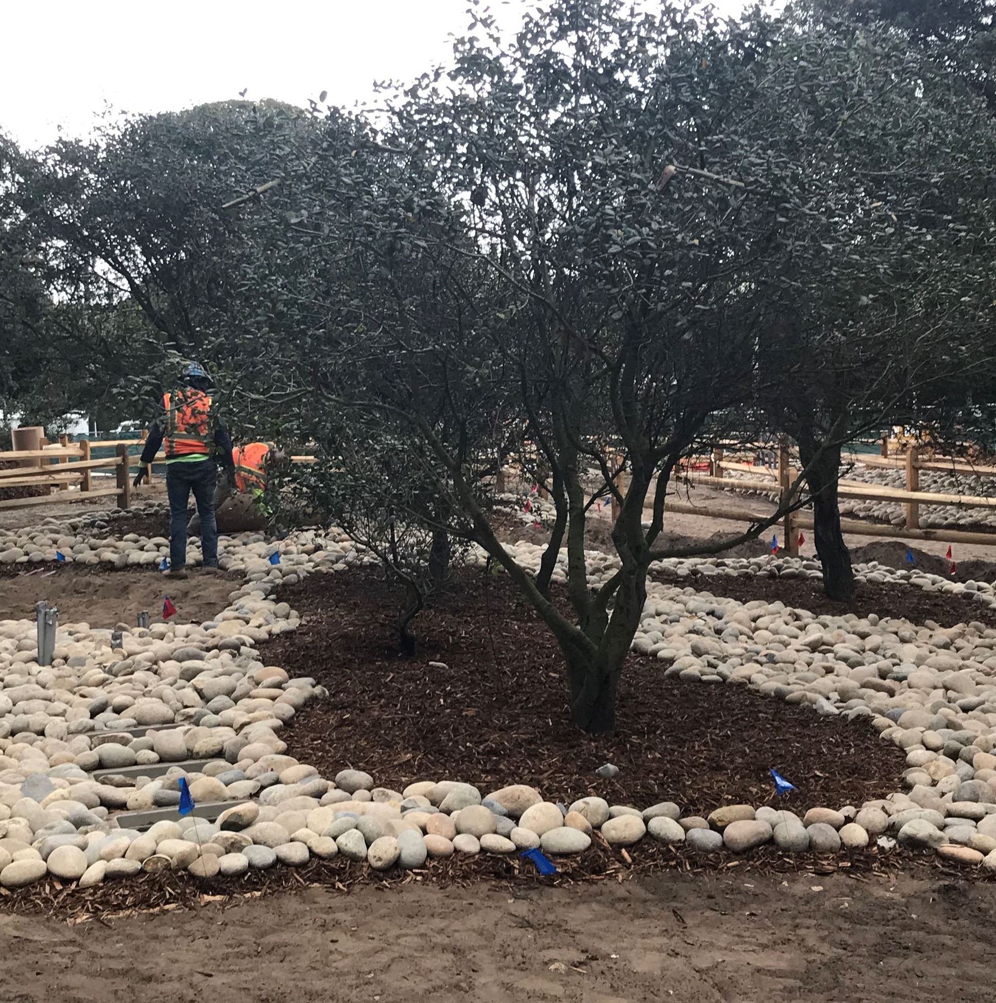 Image shows two construction workers wearing orange safety vests installing river rocks surrounding 
