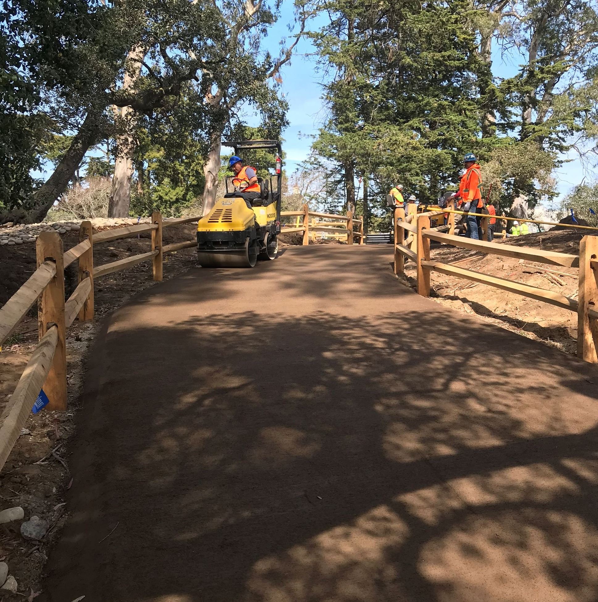 Two workers wearing orange safety vests and blue hard hats stand outside the wood split rail fence w
