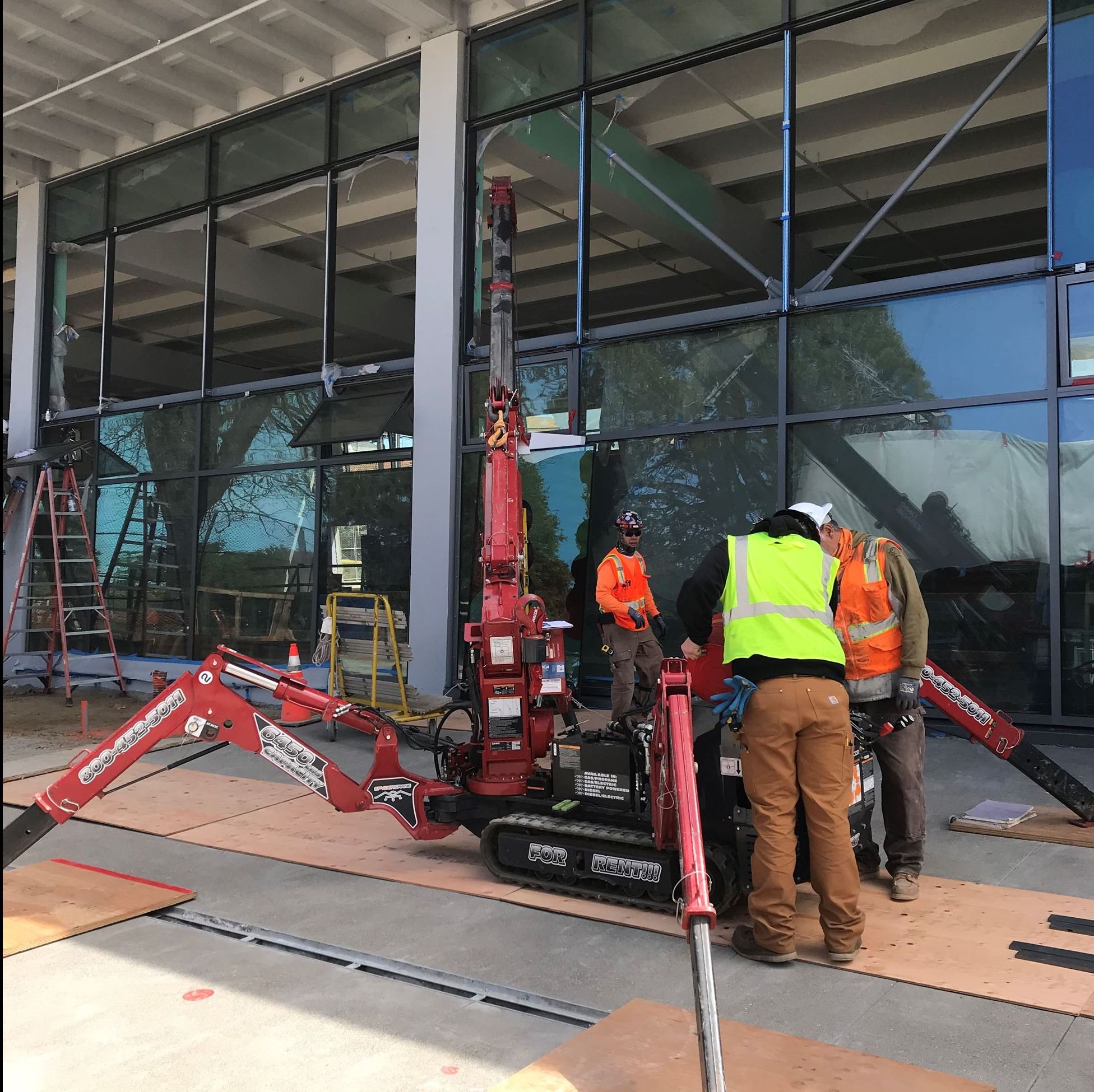 Image shows three construction workers wearing orange and yellow safety vests surrounding a machine,