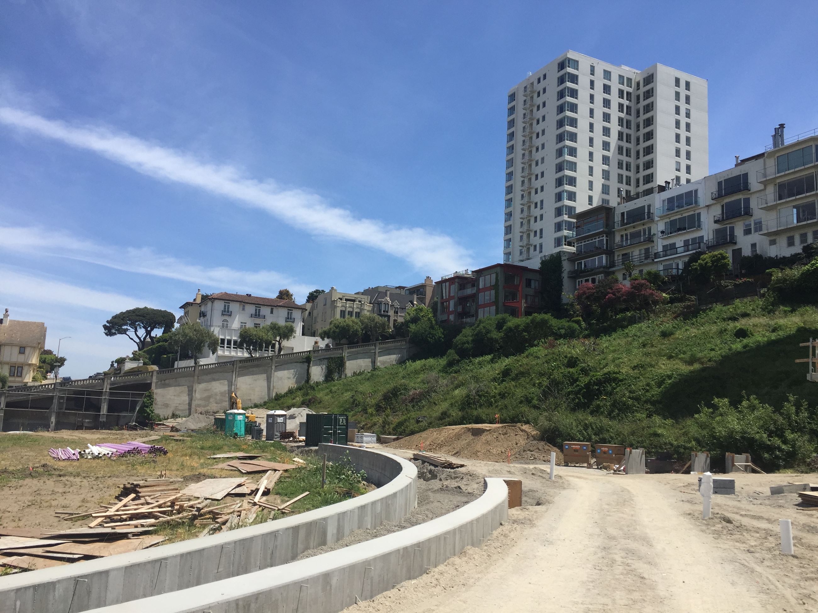 Image shows open ground with tan dirt exposed next to a large cement rounded retaining wall. Seen to