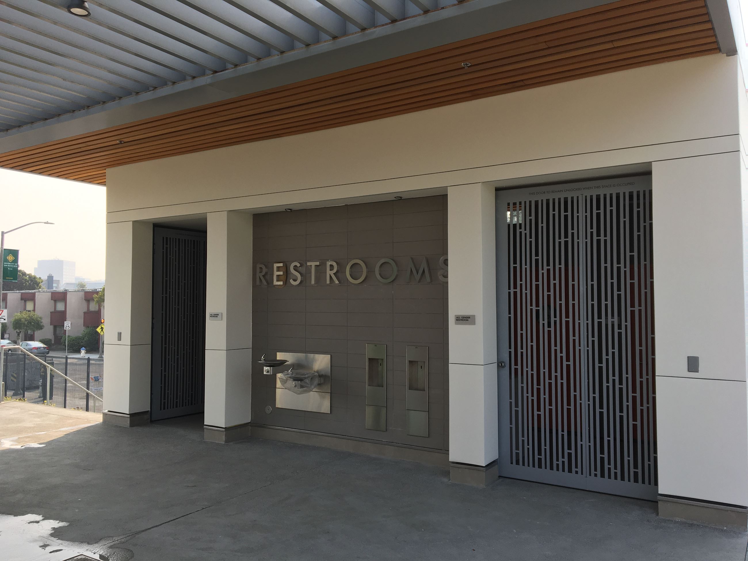 Exterior of all-gender restroom, water fountains and trash cans, metal gate closer