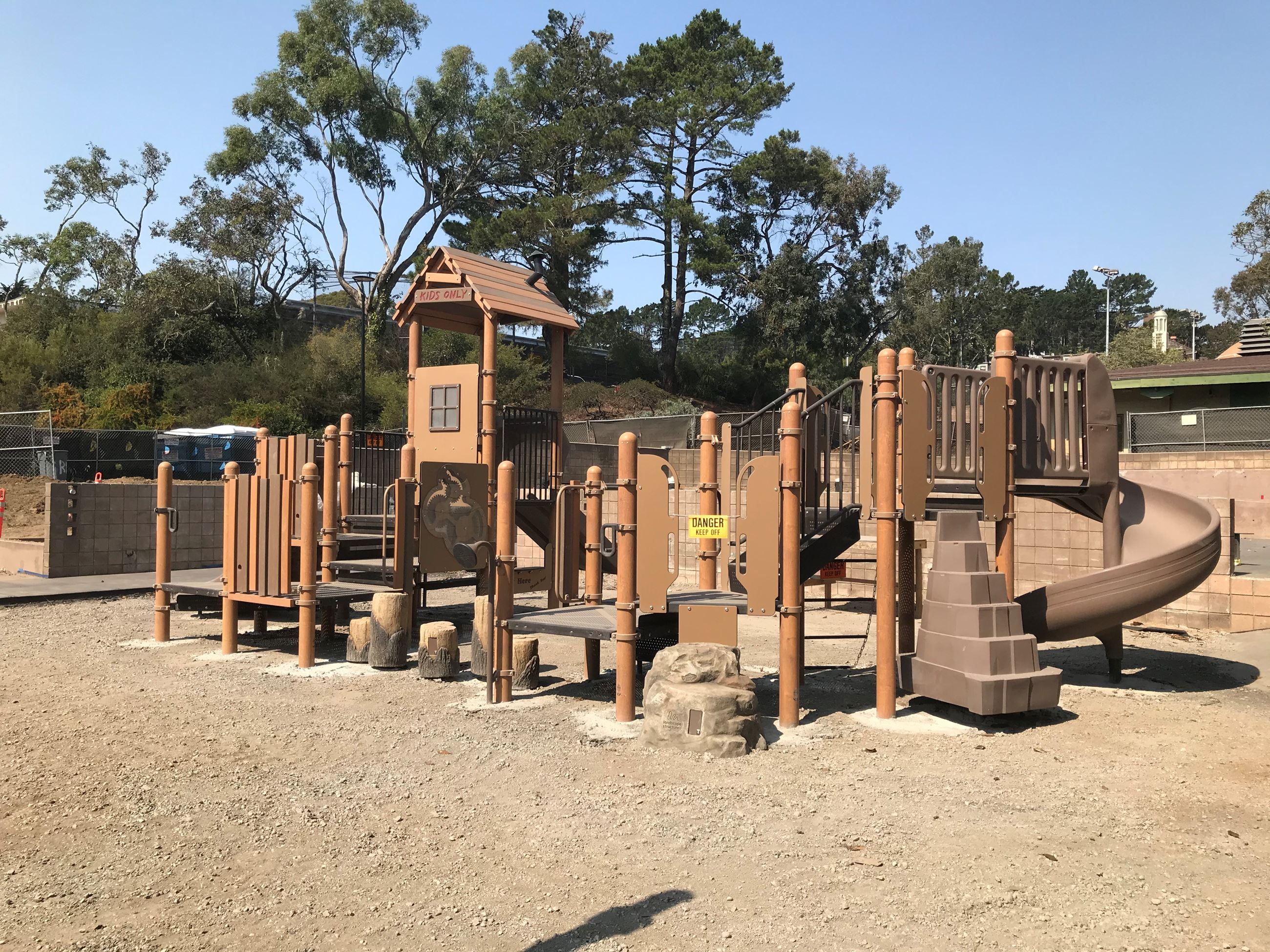 Play structure with brown climbing elements, rocks and boulders. Exposed soil.