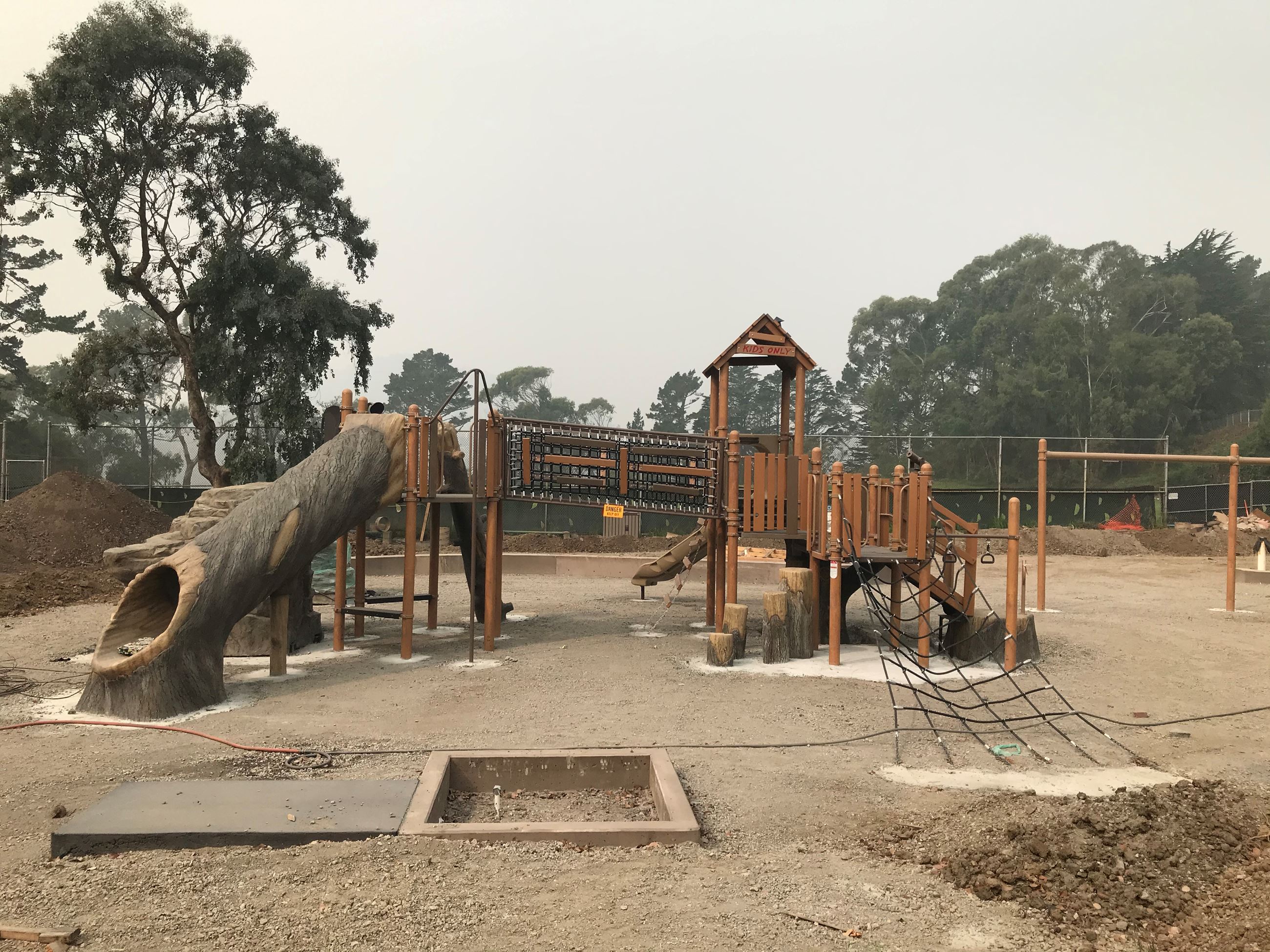 Play structure with hollow tree slide, brown bridge, exposed soil