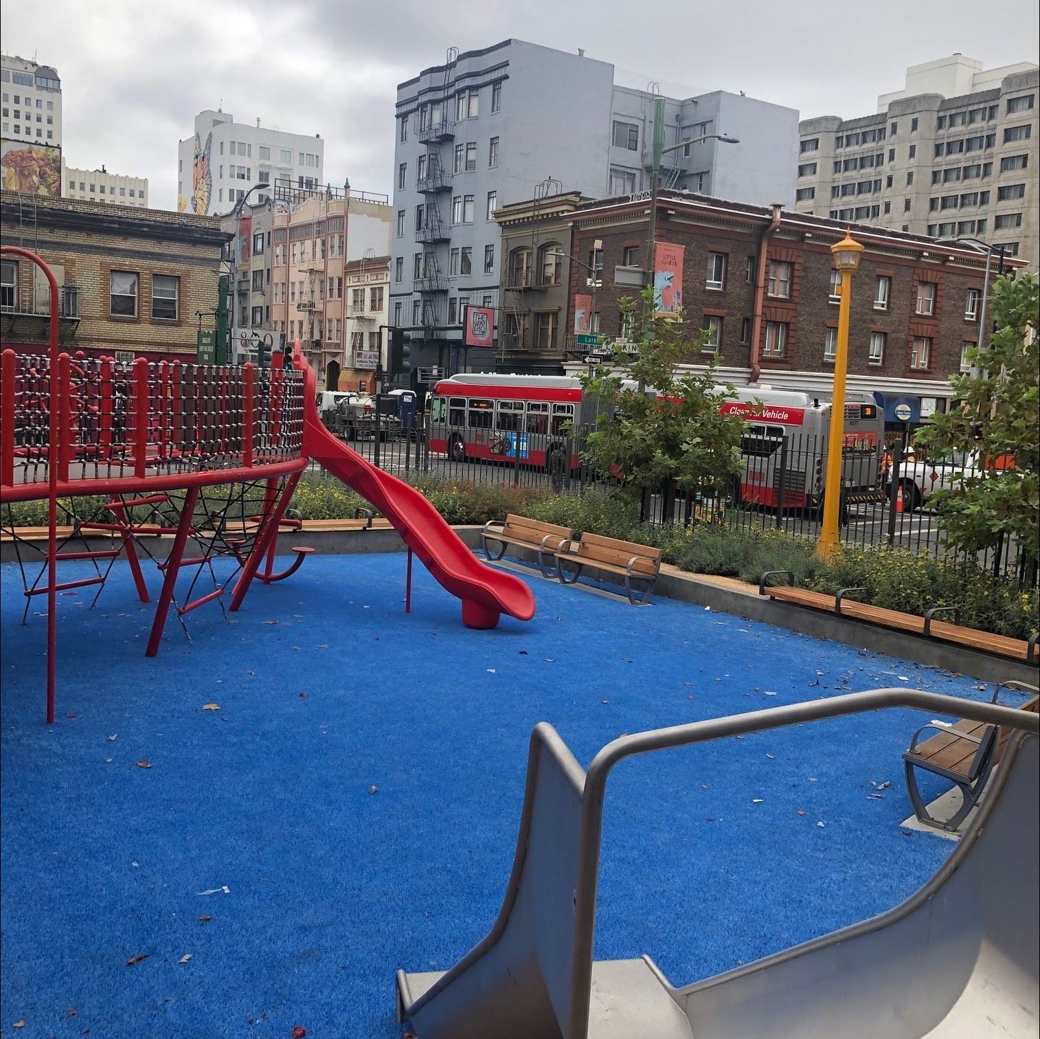 View from top of small slide, red play structure blue safety surface. buildings, bus.