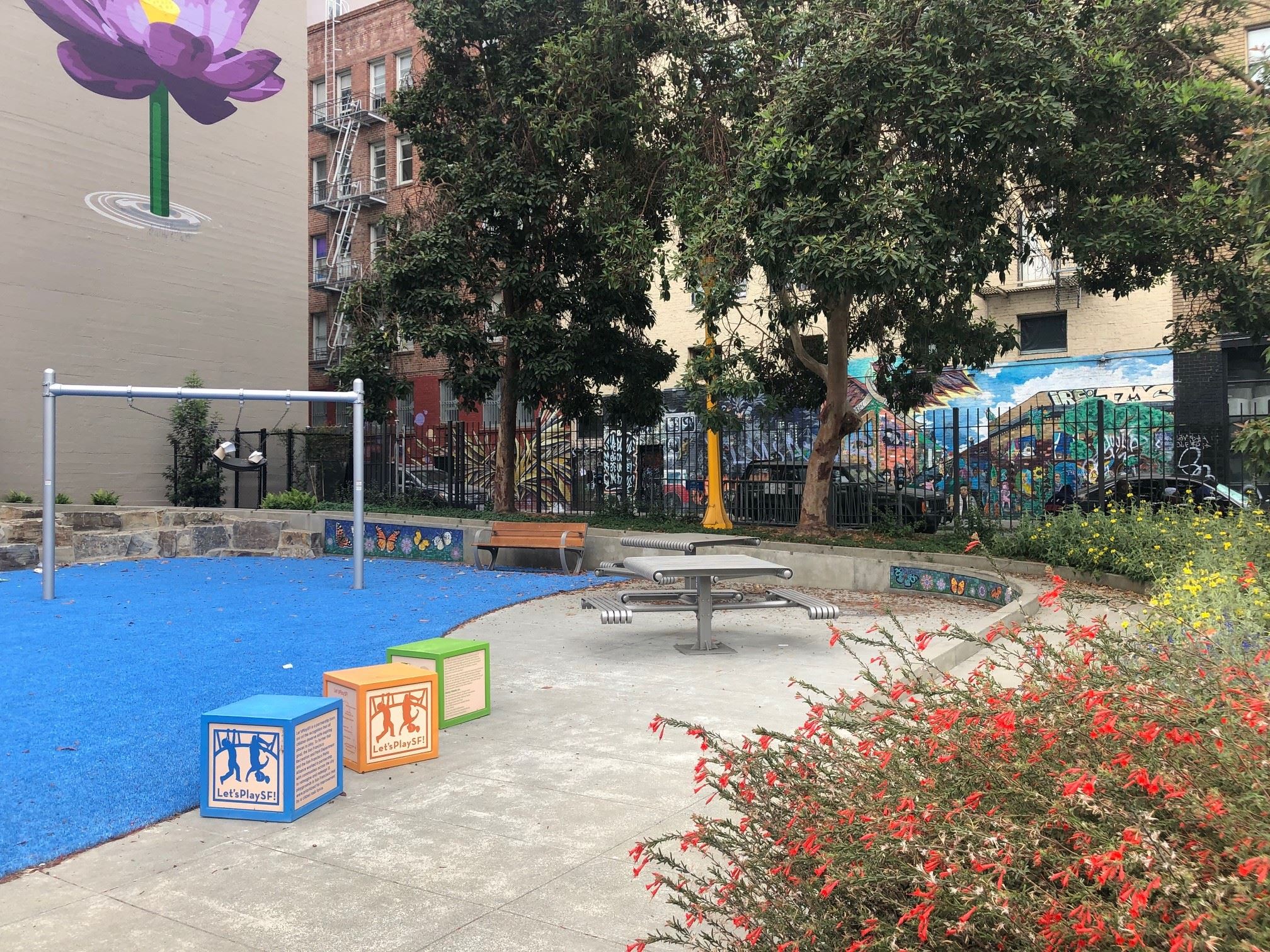 Blue play surface, metal posts with swing, purple flower painted on building, play blocks
