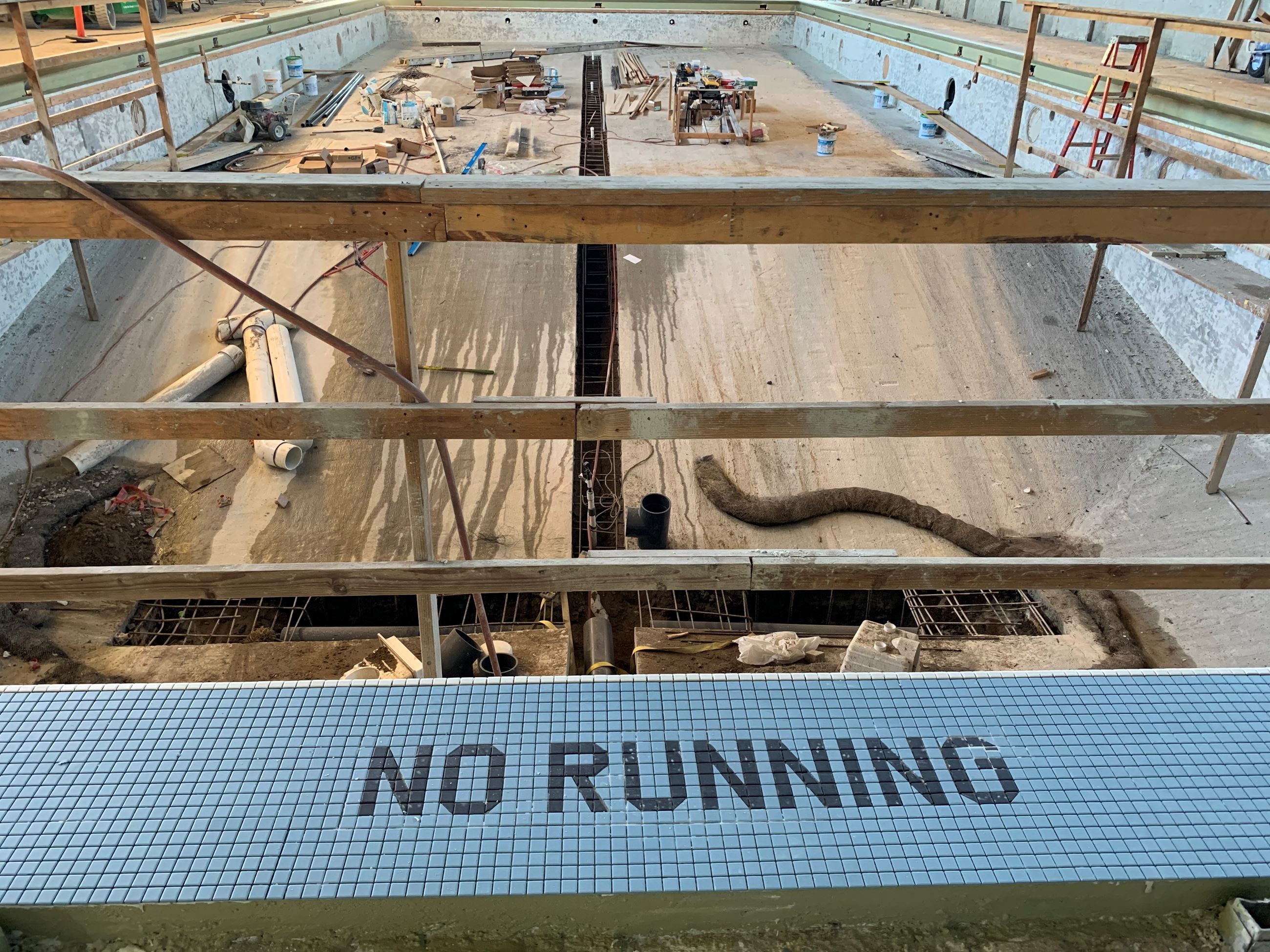 Wood temporary safety railing on the exterior of the pool. Pool deck tile with the words No Running