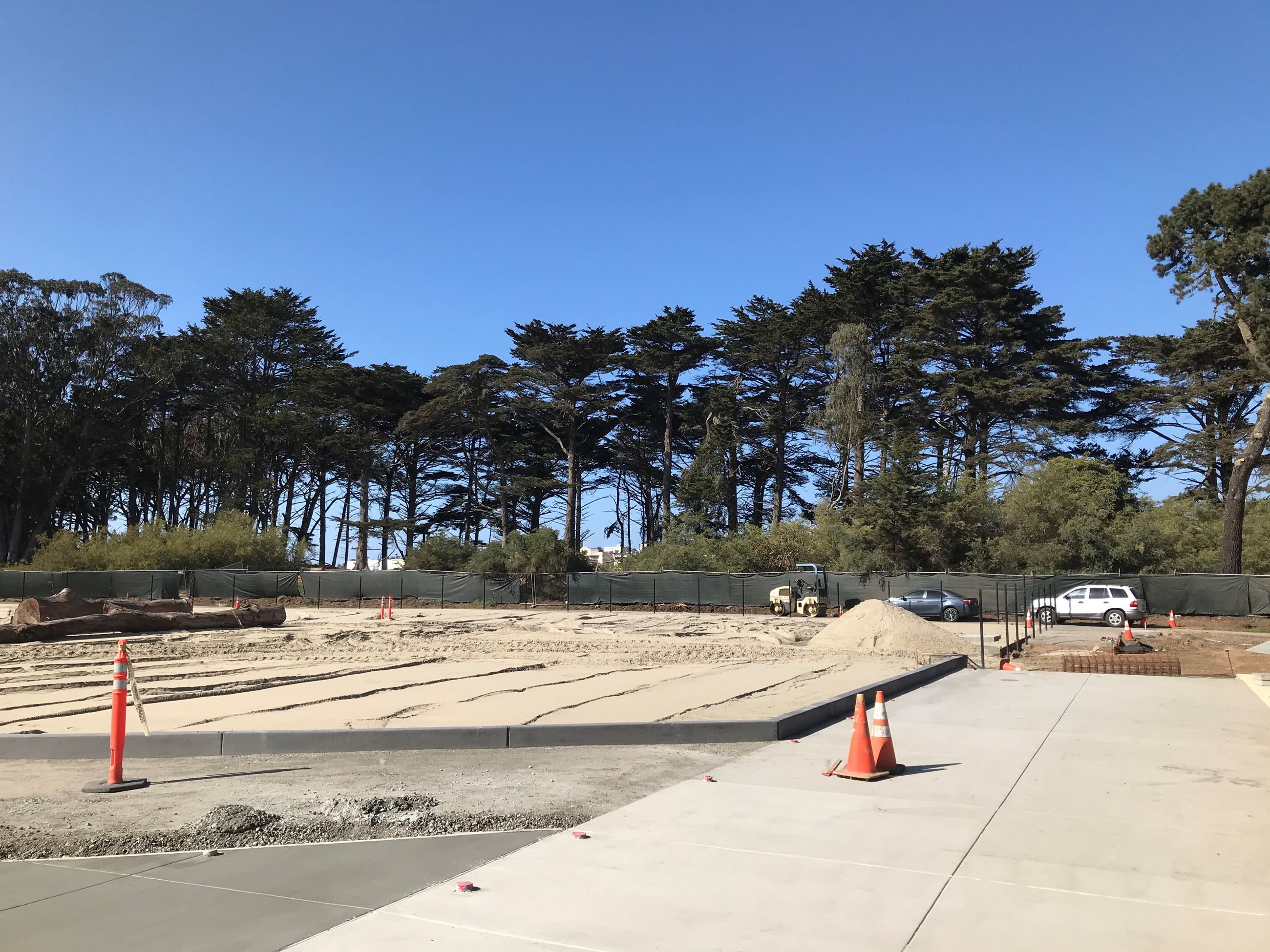 Fresh concrete poured with curb separating sand, black fence poles, two cars against green fence