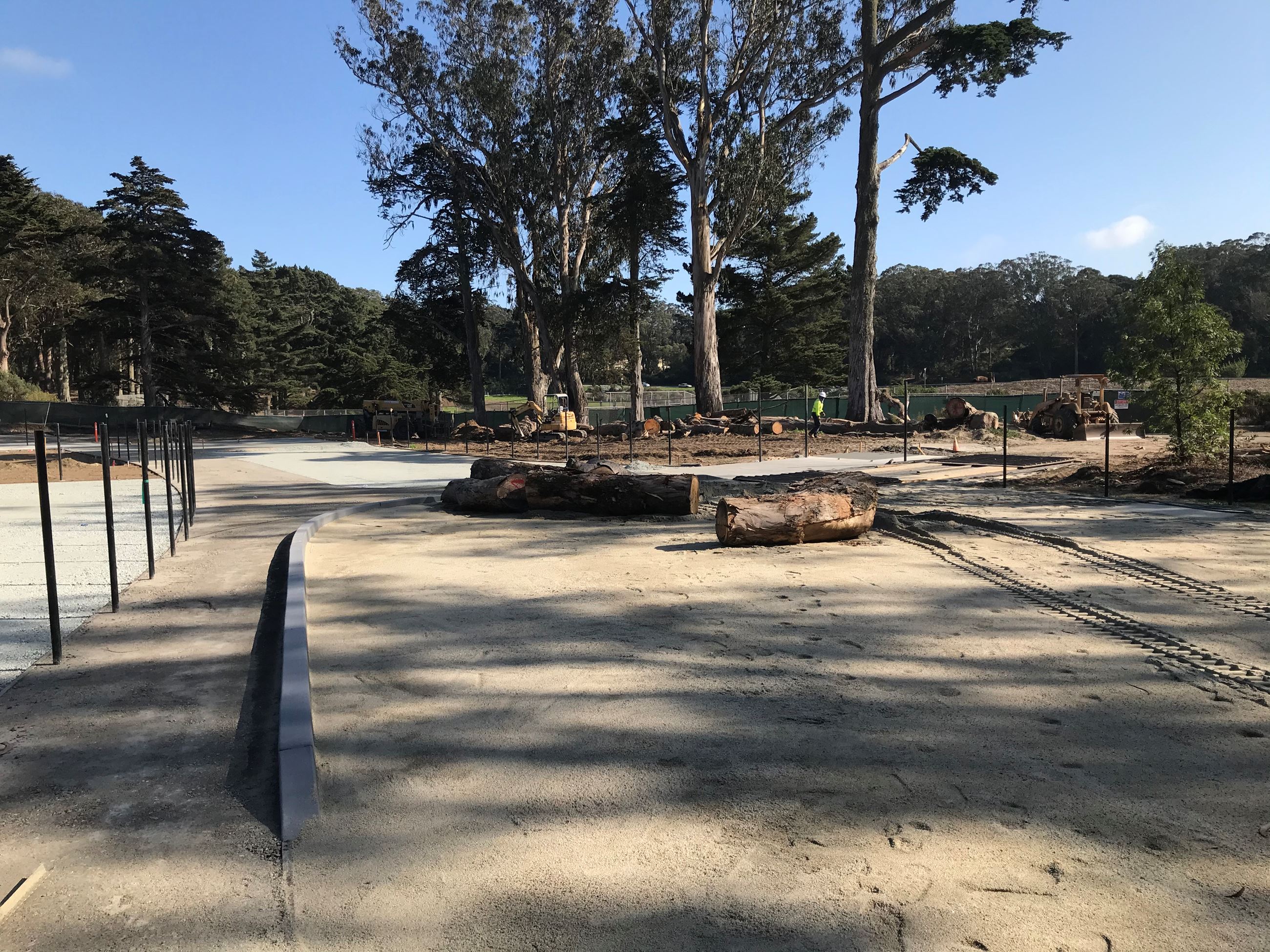 Black fence poles, cement path and curb, fresh sand with two large logs, multiple logs in background