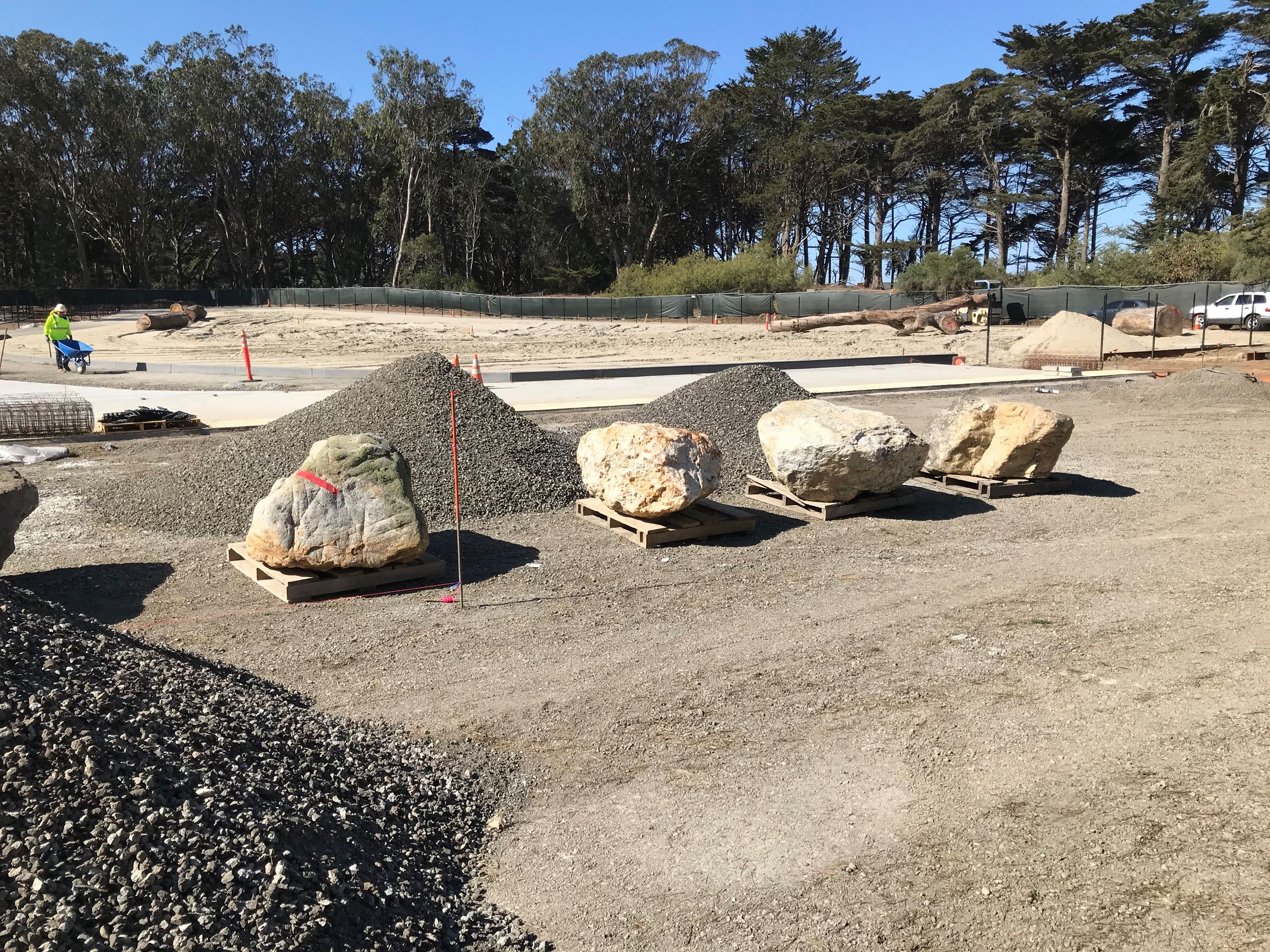 Large boulders on pallets, fresh pile of gravel, construction worker with wheelbarrow on fresh sand