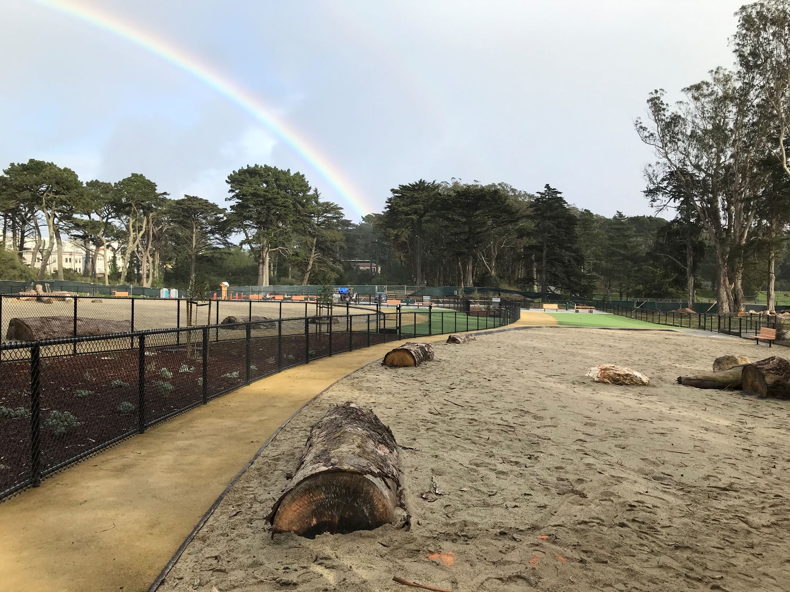 Play element giant tree trunks in sand. granitecrete pathway. Black chain link fence on the left..