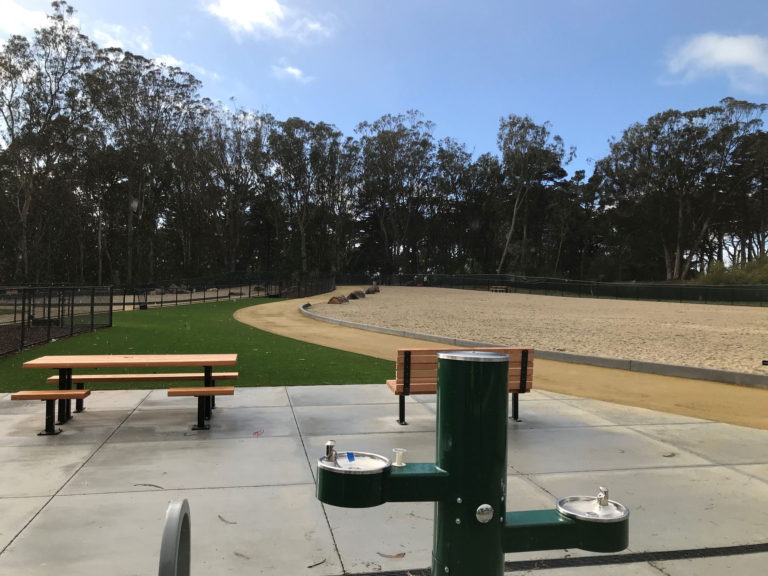 Green water fountain for all, bench and table on top of concrete pad, grass zone, pathway play area