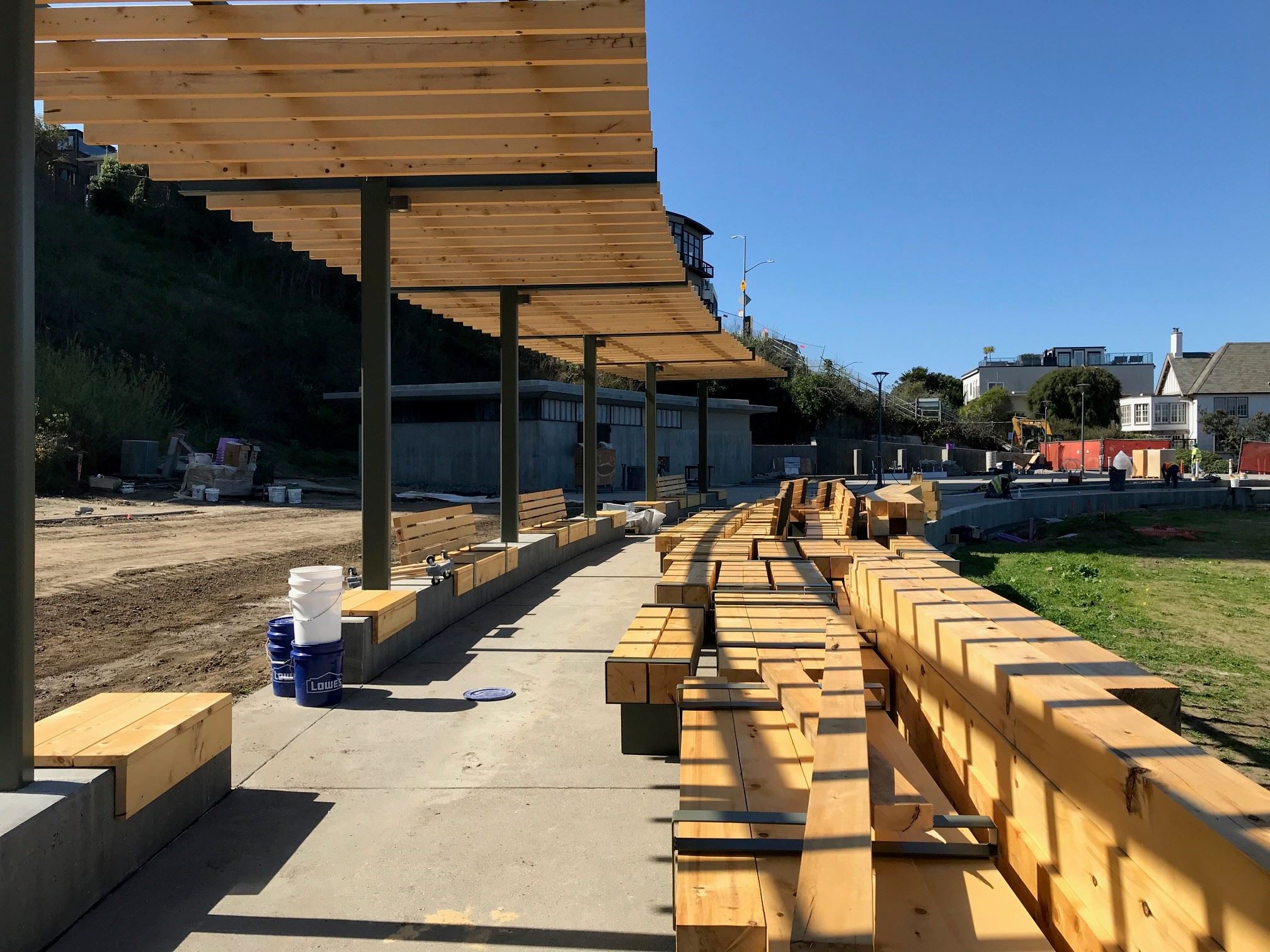 Wood slat and metal pole shade structure over sidewalk and wood benches awaiting installation