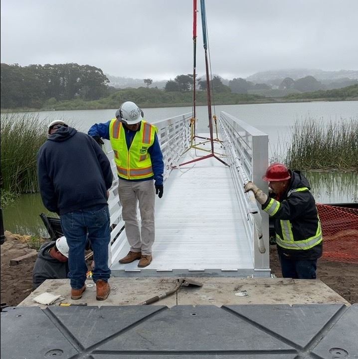Crane lowering metal gangway into place with construction workers assisting.
