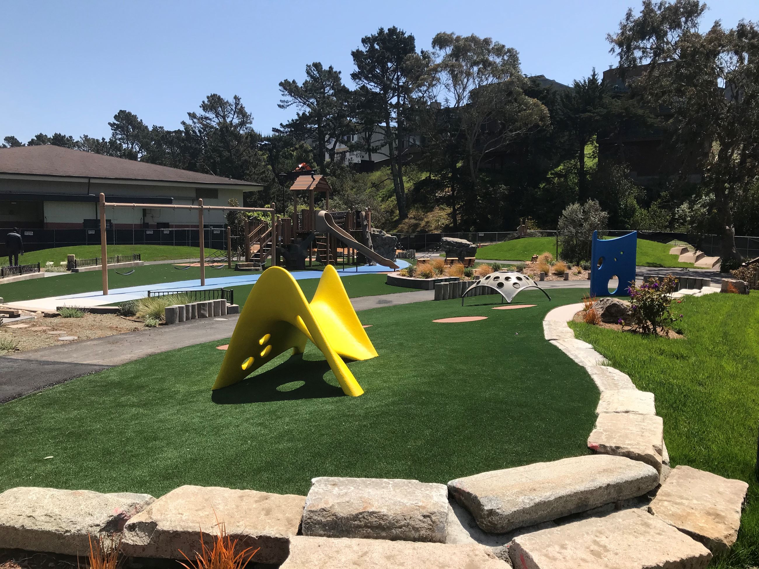 Yellow, blue and silver play structures on green safety surfacing with large wood structure behind.