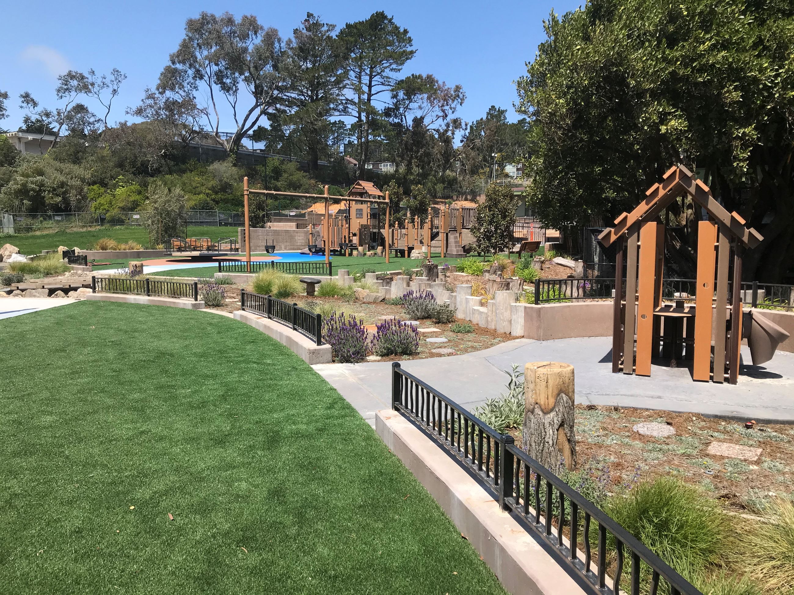 Wood slat play house surrounded by plants and natural wood elements. Small black fence on concrete.