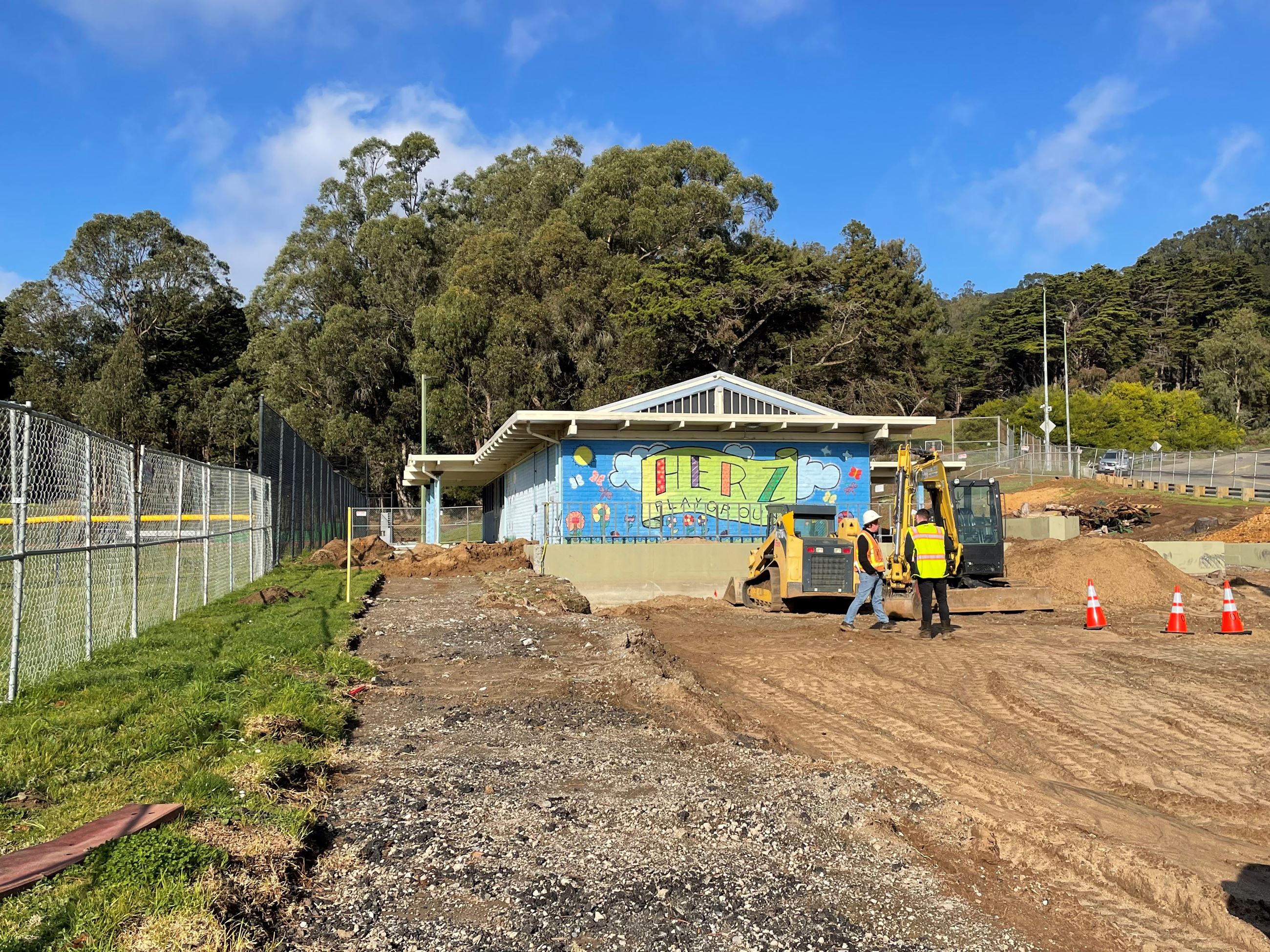 Two people wearing bright yellow standing near heavy equipment on top pf dirt exposed from demol