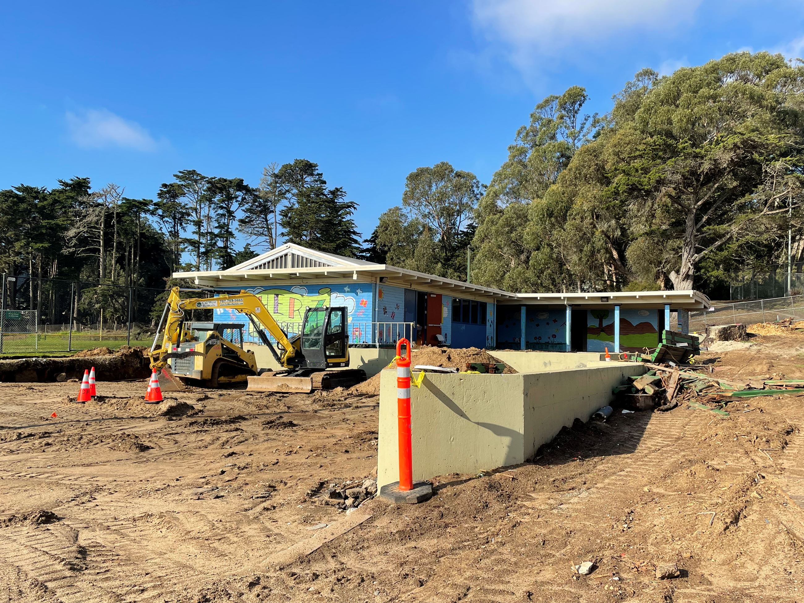 Blue clubhouse in background with exposed soil, beige short wall and heavy equipment.